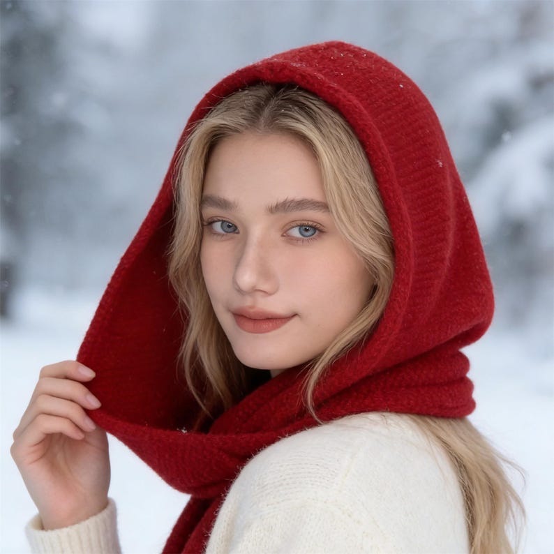 May include: A red knit hood and scarf set. The hood is pulled up, framing the face. The scarf is wrapped around the neck. The person is wearing a white sweater. The background is a snowy, blurred landscape.