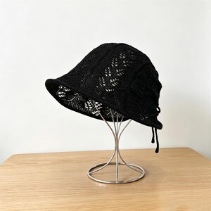 May include: A black bucket hat featuring an intricate, openwork knit pattern with leaf-like motifs. The hat has a downward-sloping brim and two thin, dangling black strings attached to the back, displayed on a silver-toned metal stand.