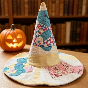 May include: A conical fabric hat featuring a patchwork design with blue, pink, and yellow sections. The hat displays various patterns including polka dots and stitched details. Beside the hat sits a carved orange pumpkin with a glowing face.