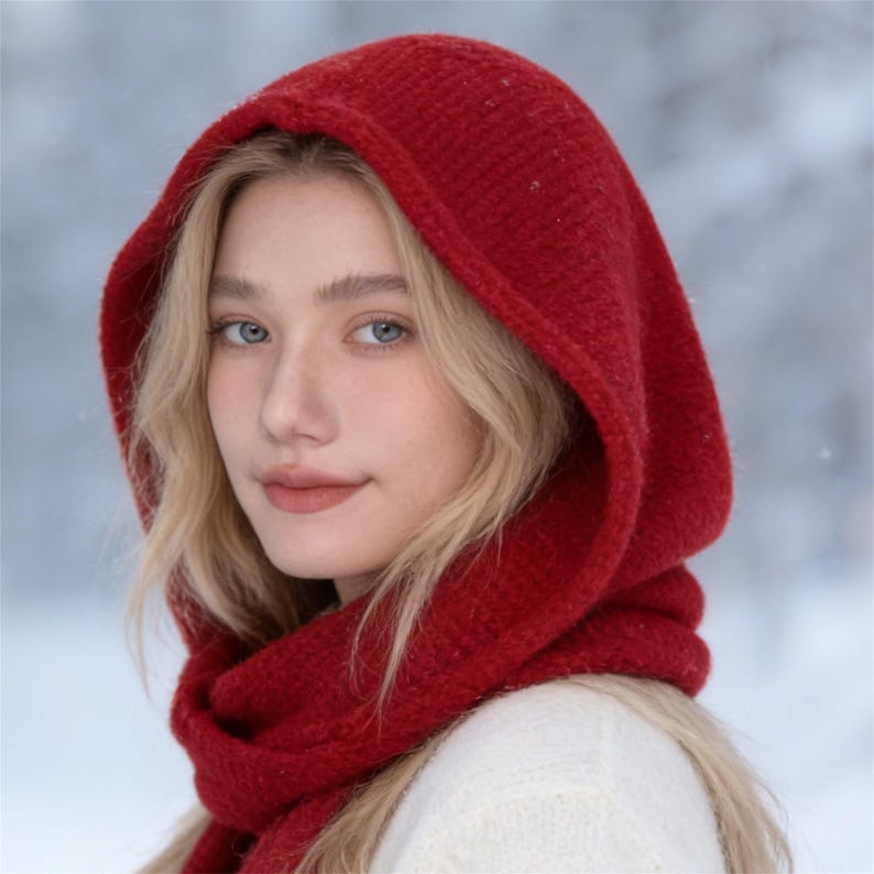 May include: A red hooded scarf, worn by a person with blonde hair and blue eyes, is the focal point of this image. The scarf is made of a thick, textured material, and the hood is pulled up over the head. The background is a snowy, blurred landscape.