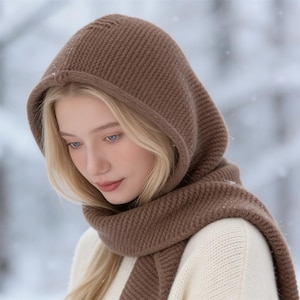May include: A brown knitted hood and scarf set. The hood frames the face and is connected to a matching scarf. The set is made of a thick, textured knit material. The background is a blurred winter scene with falling snow.