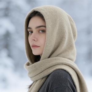 Könnte beinhalten: Ein beigefarbener Kapuzenschal aus weichem, strukturiertem Strickstoff. Die Kapuze bedeckt den Kopf und die langen Enden des Schals sind um den Hals gewickelt, was für einen Lagen-Look sorgt. Das Material hat eine flauschige, wollartige Optik.