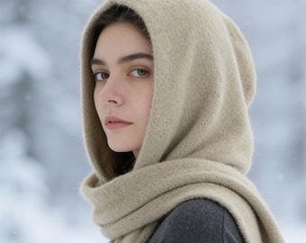 May include: A beige hooded scarf made of a soft, textured knit fabric. The hood covers the head, and the long ends of the scarf wrap around the neck, providing a layered look. The material has a fuzzy, wool-like appearance in a neutral tone.