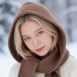 May include: A brown knitted hood and scarf set. The hood is pulled up, framing the face. The scarf is wrapped around the neck. The person is wearing a white sweater. The background is a snowy, blurred landscape.