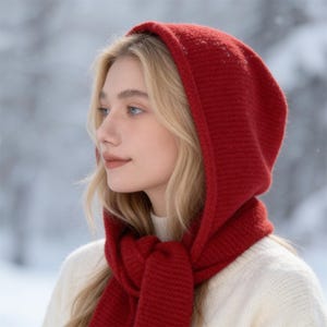 May include: A red knit hood and scarf set. The hood is pulled up, framing the face, and the scarf is wrapped around the neck. The set is made of a thick, textured knit material. The background is a snowy, winter scene.