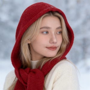 May include: A red knit hood and scarf set. The hood is pulled up, framing the face, and the scarf is draped around the neck. The woman is wearing a cream-colored sweater. The background is a snowy, blurred landscape.
