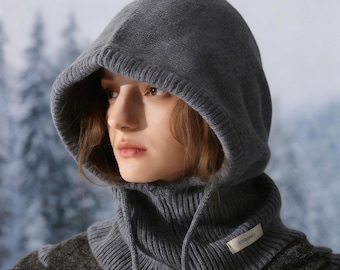 May include: A gray knit hood and cowl set. The hood features a drawstring and a ribbed knit design. The cowl is a high-neck style with a small label. The background is a blurred winter scene.