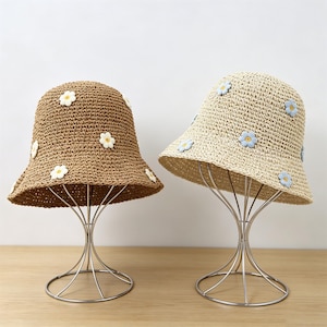May include: Two straw bucket hats, one brown and one beige, are displayed on metal stands. Each hat is adorned with small, white and blue daisy appliqués. The hats have a wide brim and a rounded crown, perfect for sun protection.