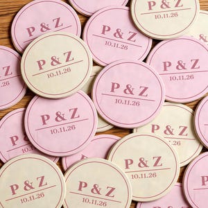 May include: A collection of round coasters in shades of light pink and cream. Each coaster features a dark pink printed design consisting of the letters P & Z above a horizontal line, with the date 10.11.26 printed directly underneath the line.