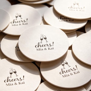 May include: A pile of circular, cream-coloured coasters. Each coaster is printed with the words "cheers!" and the names "Mia & Kai" in black, alongside a graphic of two champagne flutes. The coasters are arranged in a slightly overlapping pattern.