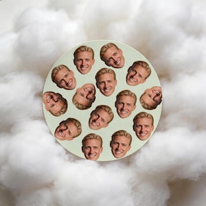 May include: A circular, pale green coaster with a repeating pattern of a smiling person's face. The faces are arranged evenly across the surface. The coaster is set against a backdrop of fluffy, white clouds.