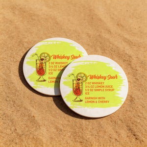 Könnte beinhalten: Zwei runde, weiße Untersetzer mit hellgrünem Hintergrund und dem Schriftzug "Whiskey Sour". Jeder Untersetzer zeigt eine Cocktail-Illustration und ein Rezept. Die Untersetzer liegen auf sandigem Untergrund.