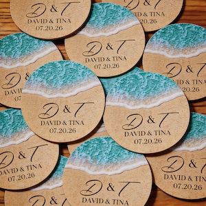 Könnte beinhalten: Eine Sammlung runder Holzuntersetzer mit einem aufgedruckten Strandmotiv, bei dem türkisfarbene Meereswellen auf beigen Sand treffen. Jeder Untersetzer ist mit dem schwarzen Text D & T, DAVID & TINA und dem Hochzeitsdatum 07.20.26 bedruckt.