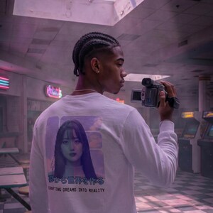 Op de afbeelding: Wit shirt met lange mouwen met een afbeelding van een vrouw en Japanse tekst. De tekst hieronder luidt "Crafting Dreams Into Reality". De persoon houdt een vintage videocamera vast in een retro arcade-omgeving.