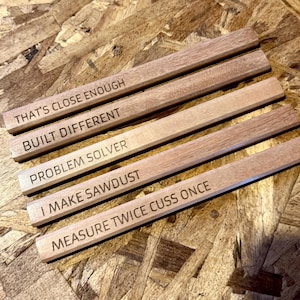 May include: Five rectangular wooden sticks with engraved text. The text reads: THAT'S CLOSE ENOUGH, BUILT DIFFERENT, PROBLEM SOLVER, I MAKE SAWDUST, and MEASURE TWICE CUSS ONCE. Each stick has a light brown wood grain finish and a smooth, flat surface.