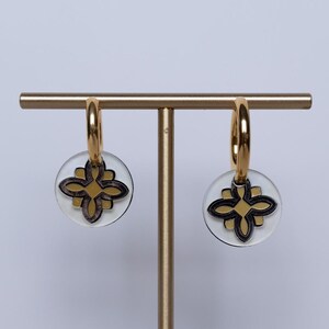 May include: Gold hoop earrings with clear, circular pendants. Each pendant has a black and gold floral design. The earrings are displayed on a gold stand against a white background.