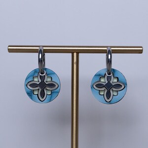 May include: A pair of circular earrings featuring a blue and black floral pattern, suspended from silver hoops. The earrings are presented on a gold-coloured stand against a white backdrop.