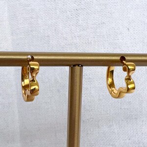 Può includere: Due orecchini a cerchio color oro con un design ondulato sono esposti su un espositore per orecchini color oro. Gli orecchini sono piccoli e hanno una finitura lucida. Lo sfondo è un tessuto bianco.