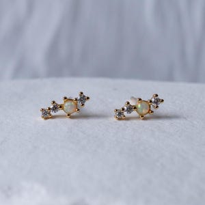 May include: A pair of gold-coloured stud earrings. Each earring has a central opal-like stone, with three smaller, clear stones either side. The earrings are on a white surface, with a soft, blurred background.