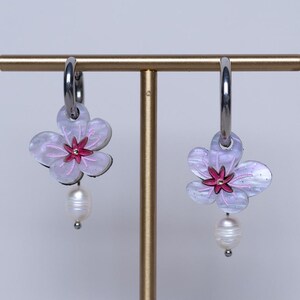 May include: Floral earrings in light pink and white. Each earring has a flower charm with a dark pink centre and a dangling white pearl. The earrings are displayed on a gold-coloured stand. The earrings are a delicate piece of jewellery.