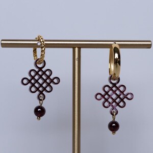 May include: A pair of gold earrings on a gold stand. One has a chain hoop, the other a smooth hoop. Both have a burgundy knot design and a small, round, burgundy bead. The earrings are displayed on a gold stand.