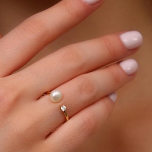 Peut inclure: Une bague ouverte en or dotée d'une perle blanche ronde à une extrémité et d'une petite pierre précieuse transparente à taille ronde à l'autre. L'anneau est fin et poli, créant un aspect délicat qui souligne le contraste entre la perle et la pierre.