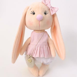 Puede incluir: Una muñeca de peluche con forma de conejo, con orejas largas, vestida con un vestido de cuadros rosas y un lazo a juego. El conejo tiene una nariz rosa, mejillas sonrosadas y un pequeño bolso con cierre de botón. La muñeca es de tela suave color melocotón claro.