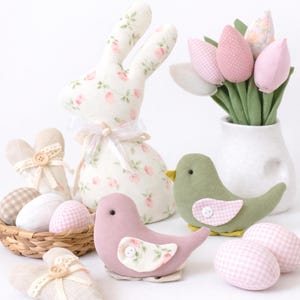 May include: An Easter display with a fabric bunny, birds, and tulips. The bunny is white with a pink floral pattern, and the birds are pink and green. Pink and white gingham eggs and a basket complete the scene. A white vase holds pink tulips.