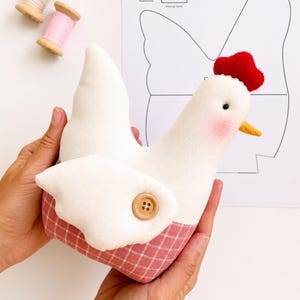 May include: A stuffed chicken toy with a white upper body, a pink and white checkered lower body, a red felt comb, a yellow beak, and a wooden button on its wing. Beside it are two wooden spools of thread and a paper sewing pattern sheet.