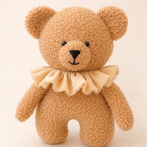 May include: A tan teddy bear made of textured, curly fabric featuring a round head, small ears, and a black embroidered nose and mouth. The bear wears a light beige, ruffled fabric collar around its neck and has two small black bead eyes.