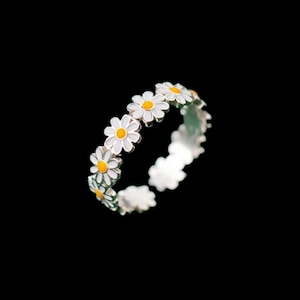 Puede incluir: Un anillo plateado con un diseño de flores de margarita. Cada margarita tiene pétalos blancos y un centro amarillo. El anillo está sobre un fondo negro.