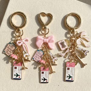 Puede incluir: Tres llaveros dorados con dijes temáticos de viaje. Cada llavero presenta un billete, la Torre Eiffel, un avión y otros dijes relacionados con viajes. Un llavero tiene un lazo rosa, otro un cierre en forma de corazón.