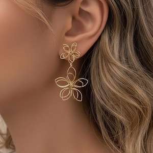 Op de afbeelding: Goudkleurige draad oorbellen in een bloemmotief. De oorbellen hebben een kleine bloemenstud en een grotere, bungelende bloem. De oorbellen worden gedragen op het oor van een persoon, met de focus op de sieraden.
