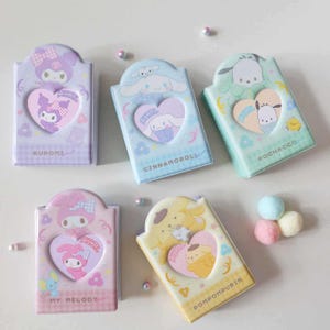 Peut inclure: Cinq boîtes décoratives rectangulaires aux couleurs pastel, chacune avec une découpe en forme de cœur. Elles présentent des personnages de dessins animés en violet, bleu clair, vert, rose et jaune. Des détails floraux et de personnages ornent les boîtes.