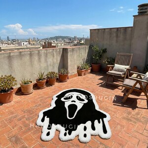 May include: A black and white Ghostface rug with a dripping design, placed on a brick patio. The rug features the iconic mask from the Scream movie franchise. The patio has potted plants and wooden chairs, with a city skyline in the background.