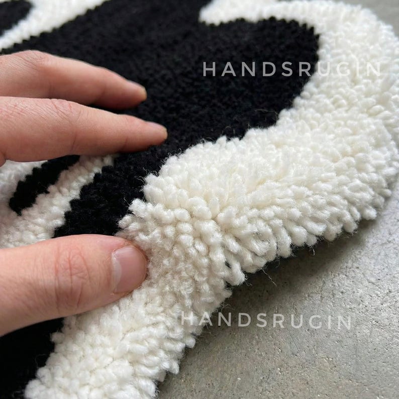 May include: Close-up of a handmade rug with a black and white pattern. The rug has a textured, looped pile. The text "HANDSRUGIN" is visible on the rug. The rug is on a gray surface.