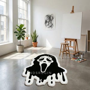 May include: A black and white rug shaped like the Ghostface mask from the Scream movie franchise. The rug has a black head and white dripping edges. The text "HANDSRUGIN" is visible on the rug.