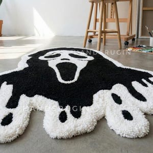 May include: A black and white tufted rug in the shape of the Ghostface mask from the Scream movie franchise. The rug features a black face with a white outline and a white background. The rug is on a gray floor.