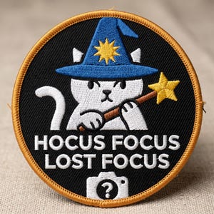 May include: Round black patch with a white cat wearing a blue wizard hat and holding a yellow star wand. Text reads "HOCUS FOCUS LOST FOCUS" with a camera icon. The patch has a gold border.