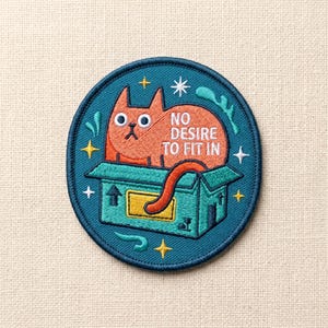 May include: Embroidered patch featuring an orange cat in a teal box, with the text "NO DESIRE TO FIT IN". The patch has a teal border with star and swirl accents. The cat has a grumpy expression.