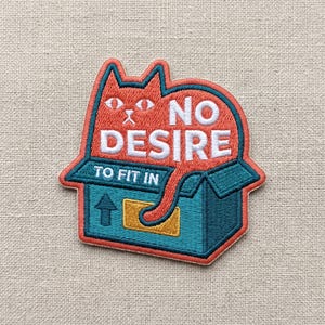 May include: Embroidered patch featuring a red cat emerging from a teal box. The patch reads "NO DESIRE TO FIT IN" in white text. The cat has orange fur and teal outlines, with a yellow accent on the box.