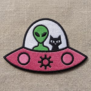 May include: Embroidered patch featuring a pink UFO with a green alien and a black cat inside. The UFO has a black outline, two pink circles, and a star-like design. The alien has large black eyes.