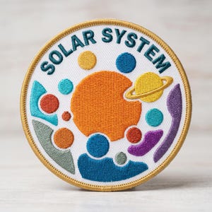 May include: Embroidered patch featuring a colorful solar system design. The patch has a white background with the words "SOLAR SYSTEM" in teal. Planets are depicted in orange, yellow, blue, and purple. The patch is round with a gold border.