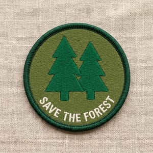 May include: A circular embroidered patch featuring two green pine trees on a green background. The words "SAVE THE FOREST" are embroidered in white along the bottom edge. The patch has a dark green border.
