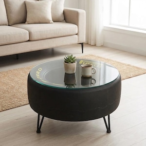 May include: A round coffee table with a black tire base and a glass top. The glass top has a yellow and green design with the word "ZERO". The table has black metal legs and a small potted plant and a mug on top.