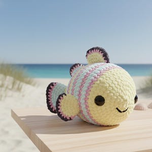 Könnte beinhalten: Ein gehäkeltes Fischspielzeug mit gelbem Körper, der mit hellblau und rosa gestreift ist. Die Flossen und die obere Flosse sind dunkelviolett mit einem gewellten Rand. Der Fisch hat schwarze Knopfaugen und ein gesticktes Lächeln. Das Spielzeug liegt auf einer Holzoberfläche mit Strand und Ozean im Hintergrund.