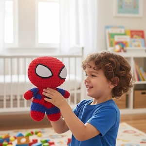 Może przedstawiać: Ręcznie robiona szydełkowa lalka Spider-Man. Lalka ma czerwoną głowę z białymi detalami oczu, niebiesko-czerwone ciało i jest trzymana przez dziecko. Tło obejmuje łóżeczko, książki i kolorowe klocki.
