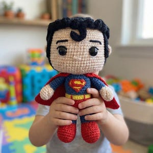 Könnte beinhalten: Handgemachte Superman-Puppe aus Häkelarbeit. Die Puppe hat schwarzes Haar, ein blau-rotes Kostüm mit dem Superman-Logo und einen roten Umhang. Die Puppe wird von einer Person gehalten.