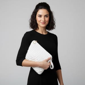 Könnte beinhalten: Eine weiße, strukturierte Clutch mit goldfarbenem Reißverschluss und Handschlaufe. Die Clutch wird vor einem schwarzen Kleid gehalten. Der Hintergrund ist einfarbig grau.