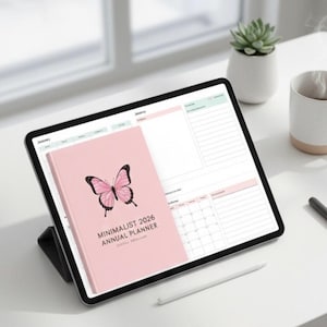 Könnte beinhalten: Ein rosa Jahresplaner 2026 mit Schmetterlingsgrafik, der auf einem Tablet mit Kalender liegt. Eine weiße Tasse, eine Sukkulente, ein Stift und ein Stift sind ebenfalls auf der weißen Oberfläche. Der Planer trägt den Titel "Minimalist 2026 Annual Planner".